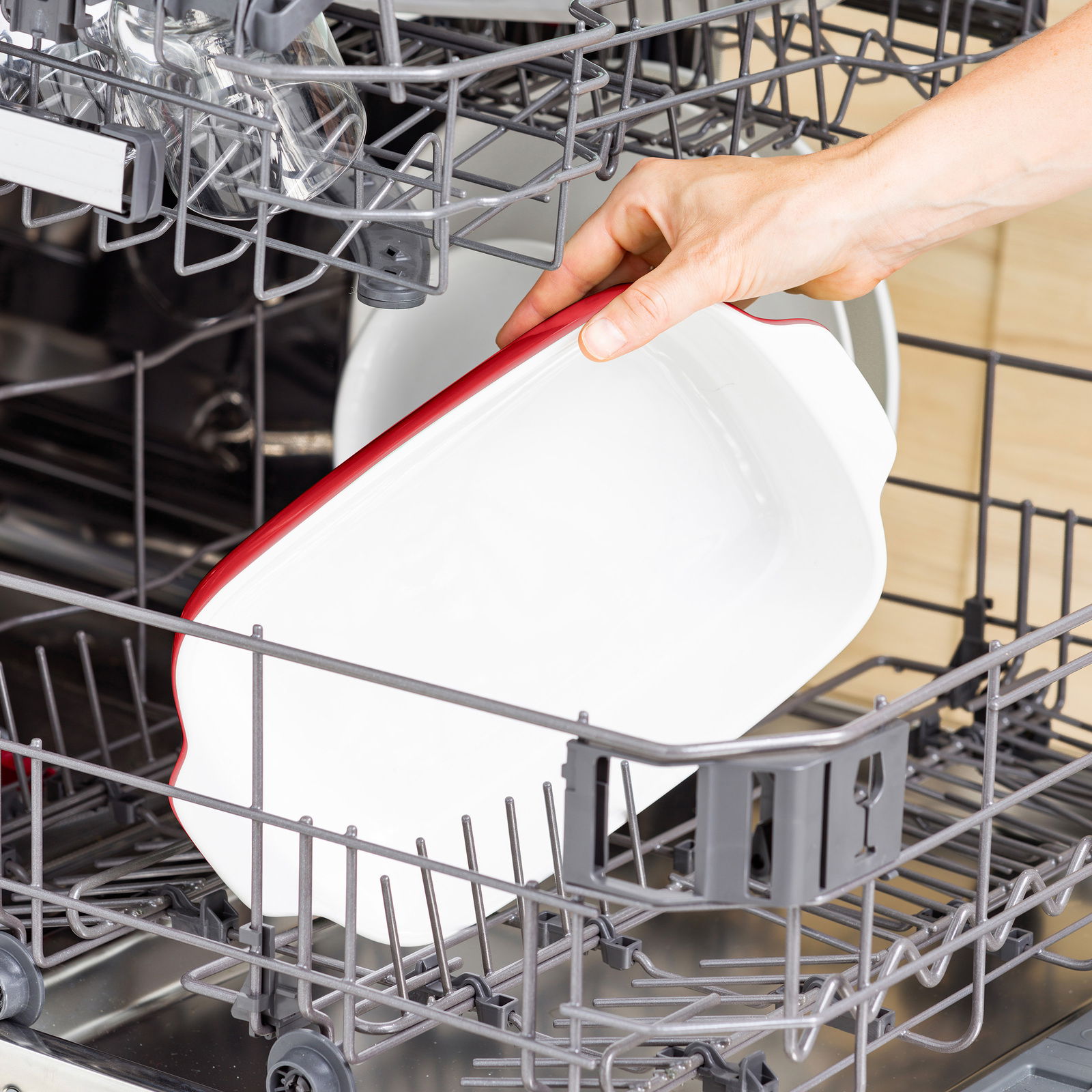 Eine weiße Auflaufform mit rotem Rand wird in eine Spülmaschine eingeräumt, seitliche Perspektive.