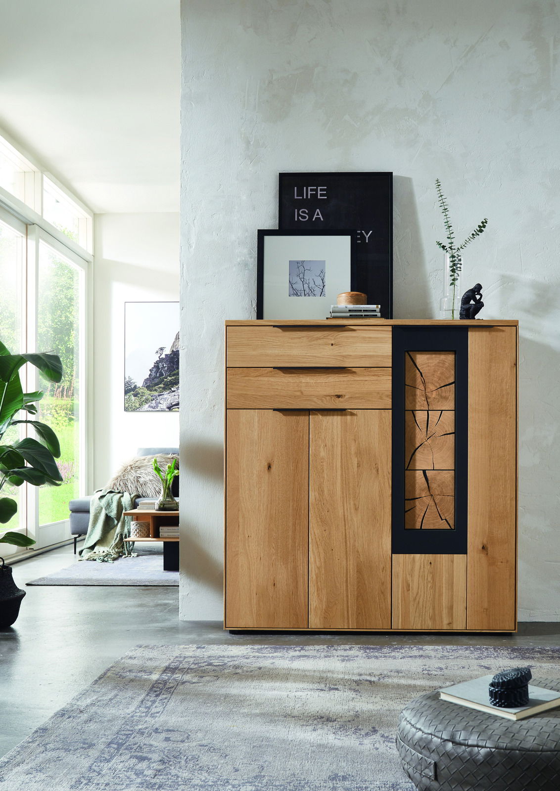 Frontale Ansicht eines modernen Highboards aus Holz mit dekorativem Glaseinsatz, umgeben von stilvoller Dekoration in einem hellen Wohnzimmer.