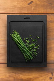 Schwarzes Schneidebrett der Marke WOLL mit geschnittenem Schnittlauch, von oben fotografiert auf einer Holzoberfläche.