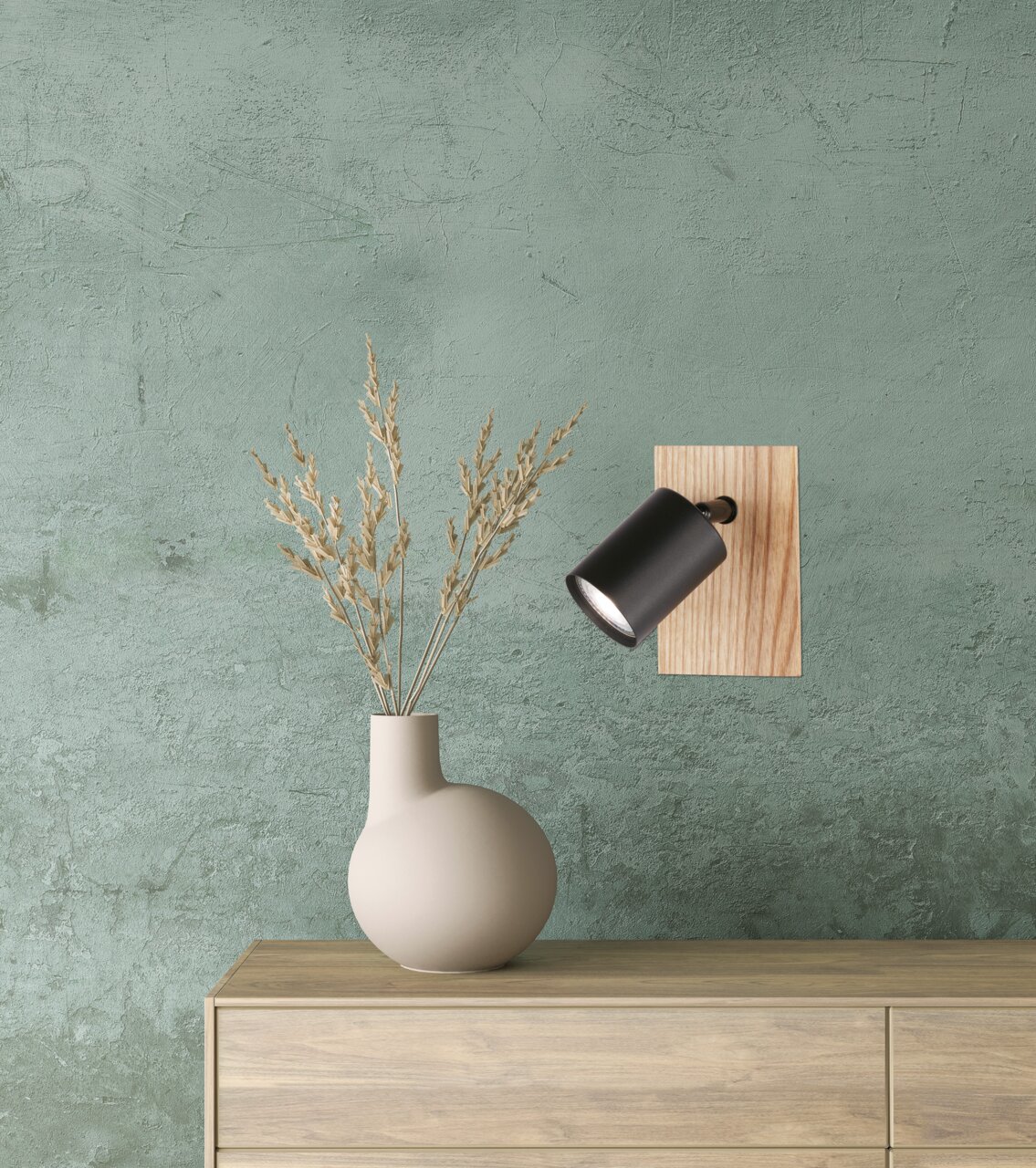 Moderne wandlamp Holton met zwarte lampenkap en houten achterwand, zijaanzicht, bevestigd aan een groene muur boven een commode met vaas.
