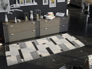 Moderner Teppich in Grau- und Schwarztönen mit geometrischem Muster, auf dunklem Boden vor einer Kommode liegend, aus der Vogelperspektive betrachtet.