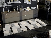 Moderner Teppich in Grau- und Schwarztönen mit geometrischem Muster, auf dunklem Boden vor einer Kommode liegend, aus der Vogelperspektive betrachtet.