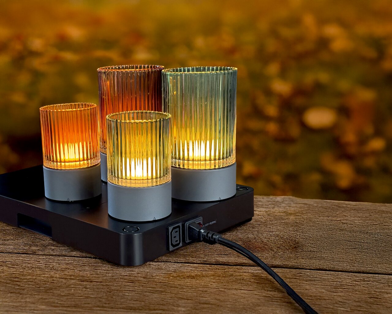 Vier bunte Akku-Windlichter auf einer Ladestation, auf einem Holztisch im Freien, mit herbstlichem Hintergrund.
