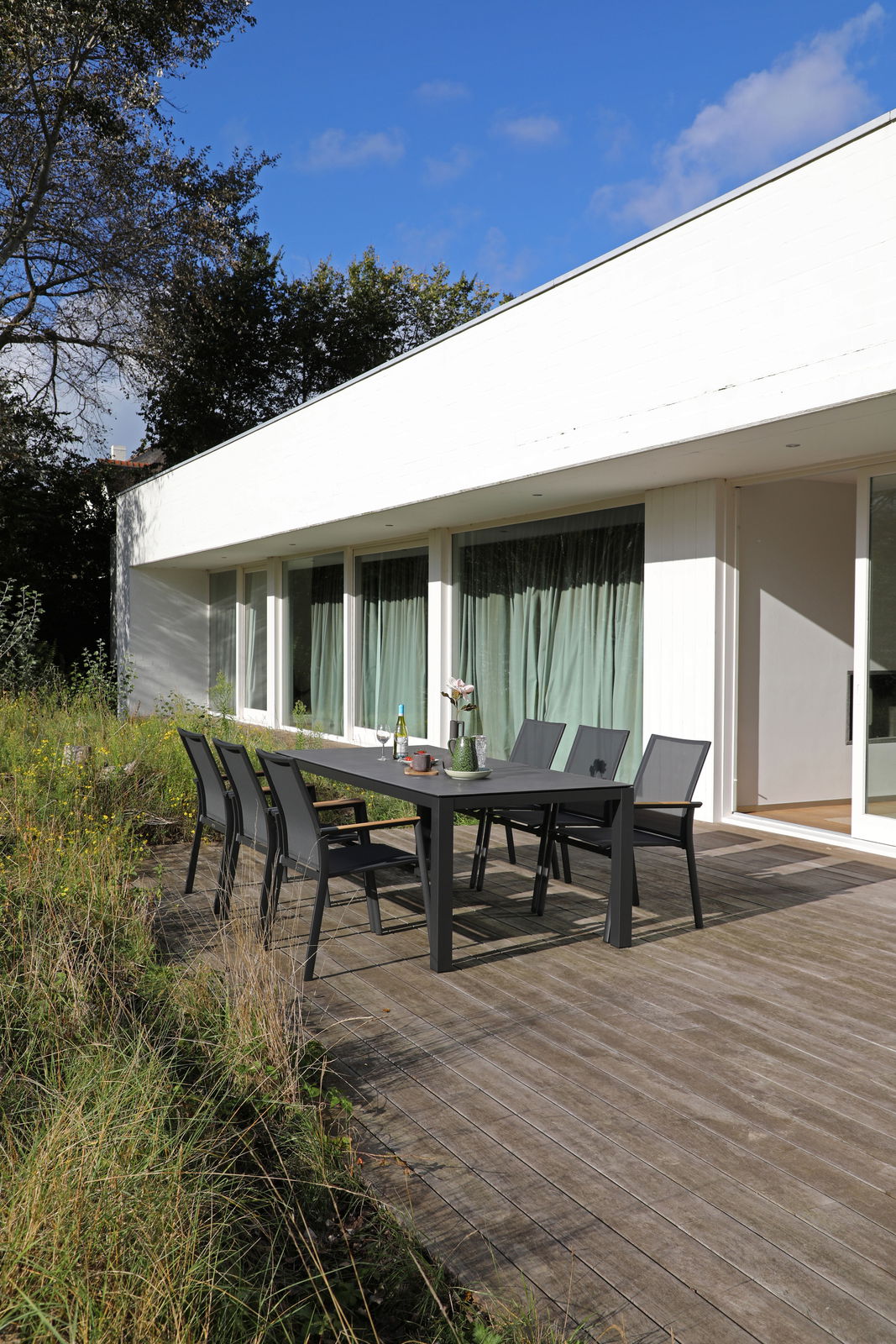Moderne Gartenmöbel auf einer Terrasse mit Blick auf ein weißes Gebäude, bestehend aus einem großen Tisch und mehreren Stapelsesseln.