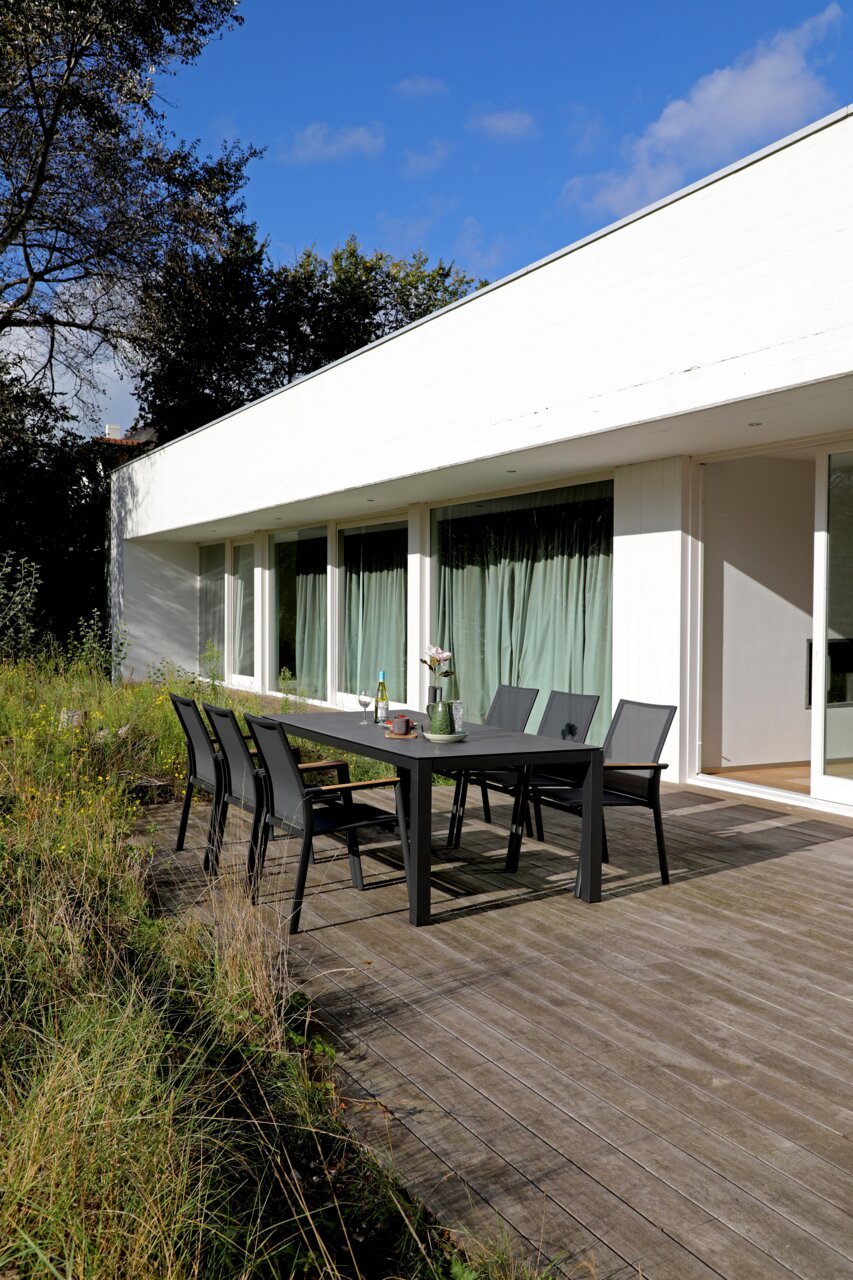 Moderne Gartenmöbel auf einer Terrasse mit Blick auf ein weißes Gebäude, bestehend aus einem großen Tisch und mehreren Stapelsesseln.