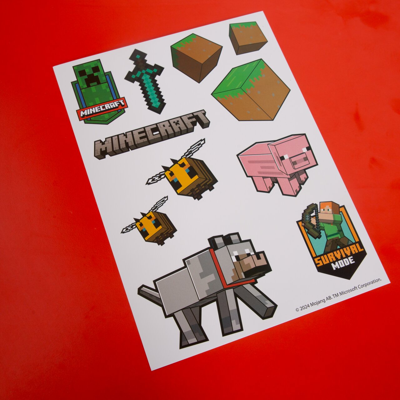 Minecraft Stickerbogen mit Creeper, Diamantschwert, Grasblöcken, Bienen, Schwein, Wolf, Minecraft-Logo und Survival-Mode-Motiv auf weißem Blatt vor rotem Hintergrund, fotografiert in schräger Draufsicht (Top-Down-Perspektive).