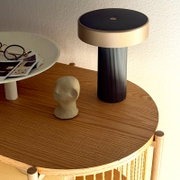 Moderne kabellose LED-Tischleuchte mit geriffeltem schwarzem Fuß und rundem beigefarbenem Schirm auf Holz-Beistelltisch, seitliche Perspektive leicht von oben