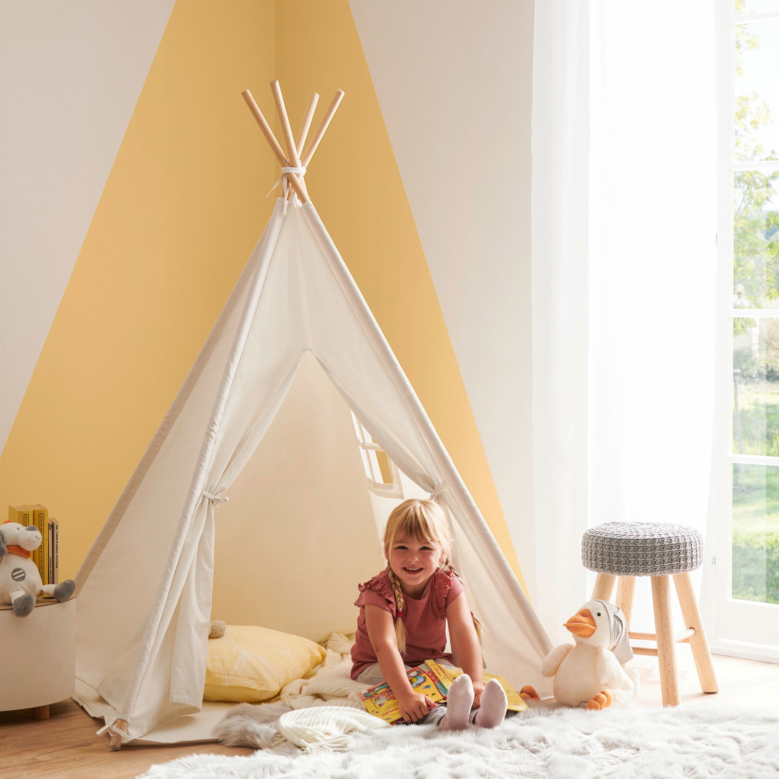 Kind sitzt lächelnd in einem weißen Spielzelt in einem hellen Raum, umgeben von Kuscheltieren und einem Hocker. Perspektive von vorne.