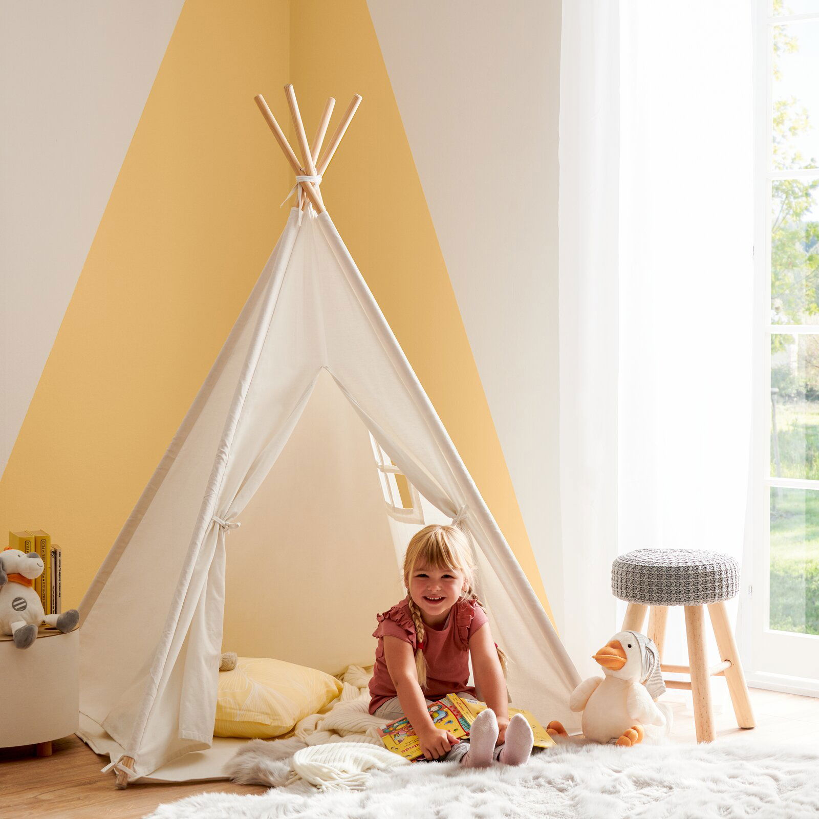 Kind sitzt lächelnd in einem weißen Spielzelt in einem hellen Raum, umgeben von Kuscheltieren und einem Hocker. Perspektive von vorne.
