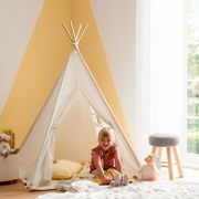 Kind sitzt lächelnd in einem weißen Spielzelt in einem hellen Raum, umgeben von Kuscheltieren und einem Hocker. Perspektive von vorne.