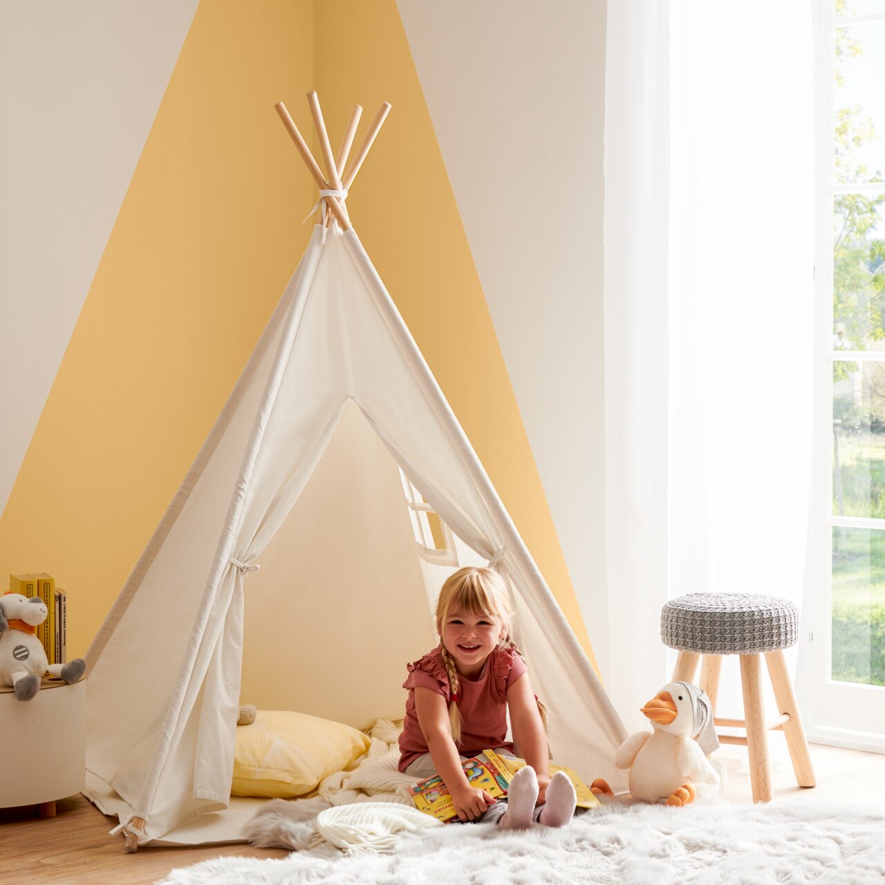 MyBaby Spielzelt JULIAN Kind sitzt lächelnd in einem weißen Spielzelt in einem hellen Raum, umgeben von Kuscheltieren und einem Hocker. Perspektive von vorne.