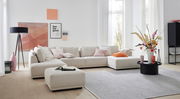 Modernes Ecksofa in hellem Beige mit Kissen in verschiedenen Farben, umgeben von stilvoller Dekoration in einem hellen Wohnzimmer. Perspektive von vorne.