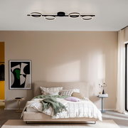 Moderne schwarze LED-Deckenleuchte mit vier ringförmigen Lichtquellen auf einer geraden Schiene, warmweiß leuchtend, montiert an der Decke über einem Schlafzimmer; Perspektive: frontale Raumansicht leicht von unten.