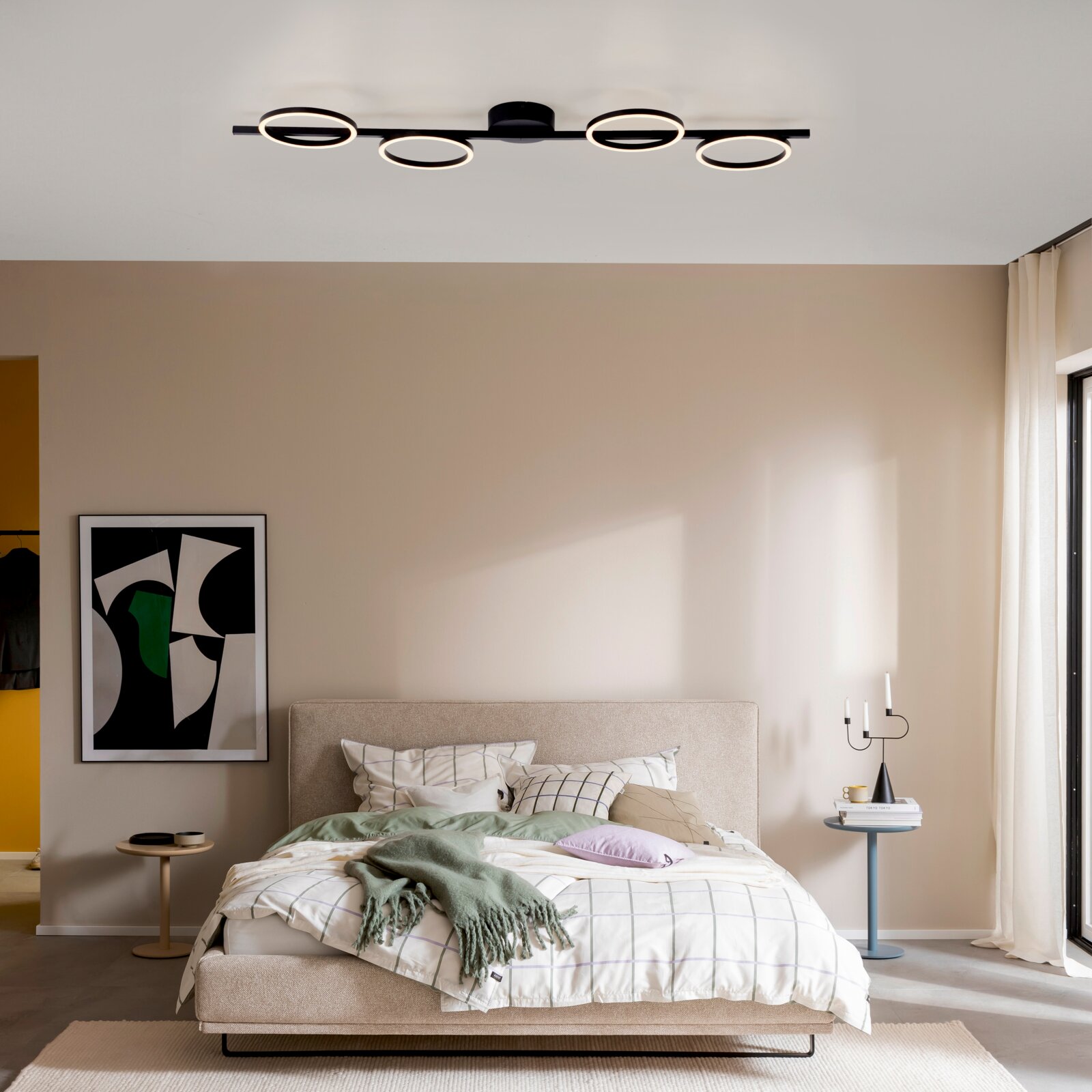 Moderne schwarze LED-Deckenleuchte mit vier ringförmigen Lichtquellen auf einer geraden Schiene, warmweiß leuchtend, montiert an der Decke über einem Schlafzimmer; Perspektive: frontale Raumansicht leicht von unten.