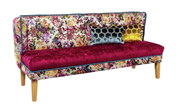 Bunt gemustertes 2-Sitzer-Sofa mit floraler Rückenlehne, bordeauxroter Sitzfläche, zwei Zierkissen (Wabenmuster in Blau und Gelb) und Holzfüßen, freigestellt auf Weiß, Ansicht schräg von vorne.