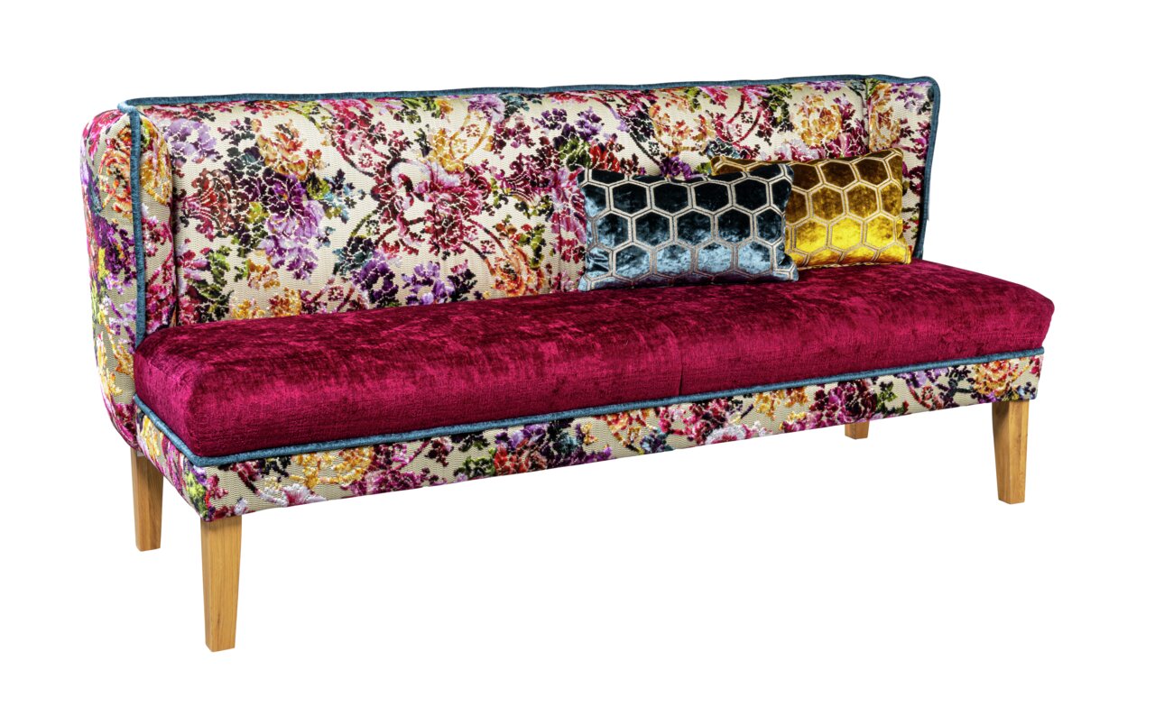 Bunt gemustertes 2-Sitzer-Sofa mit floraler Rückenlehne, bordeauxroter Sitzfläche, zwei Zierkissen (Wabenmuster in Blau und Gelb) und Holzfüßen, freigestellt auf Weiß, Ansicht schräg von vorne.