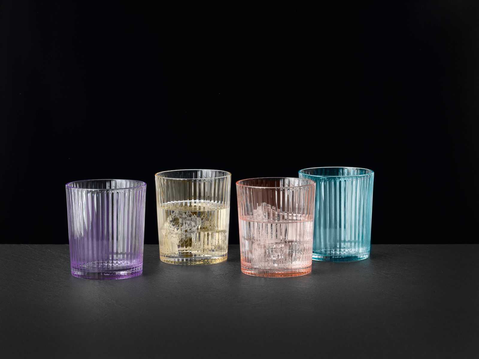 Nachtmann Becherset 4-tlg. APERITIVO Vier bunte Becher aus Glas in verschiedenen Farben (Lila, Gelb, Rosa, Blau) auf schwarzem Hintergrund, frontal betrachtet.