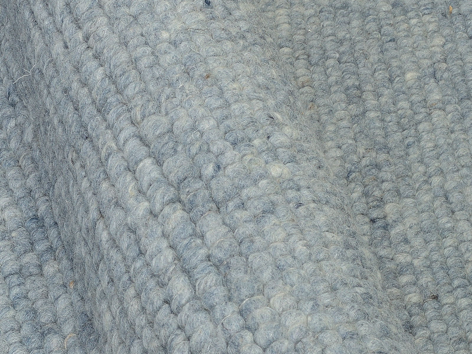 Nahaufnahme eines handgewebten Teppichs in graublauer Farbe mit strukturierter Oberfläche, seitliche Perspektive.