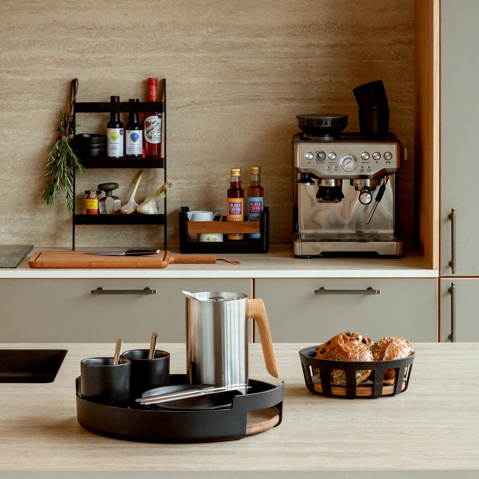 Modernes Küchen-Setup mit schwarzem Tablett-Set im Vordergrund (Becher, Schale, Besteck), Edelstahlkanne mit Holzgriff und Brotkorb; Hintergrund mit Regal und Espressomaschine, aufgenommen in frontaler Perspektive auf Arbeitsplattenhöhe.