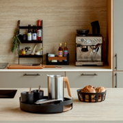 Modernes Küchen-Setup mit schwarzem Tablett-Set im Vordergrund (Becher, Schale, Besteck), Edelstahlkanne mit Holzgriff und Brotkorb; Hintergrund mit Regal und Espressomaschine, aufgenommen in frontaler Perspektive auf Arbeitsplattenhöhe.