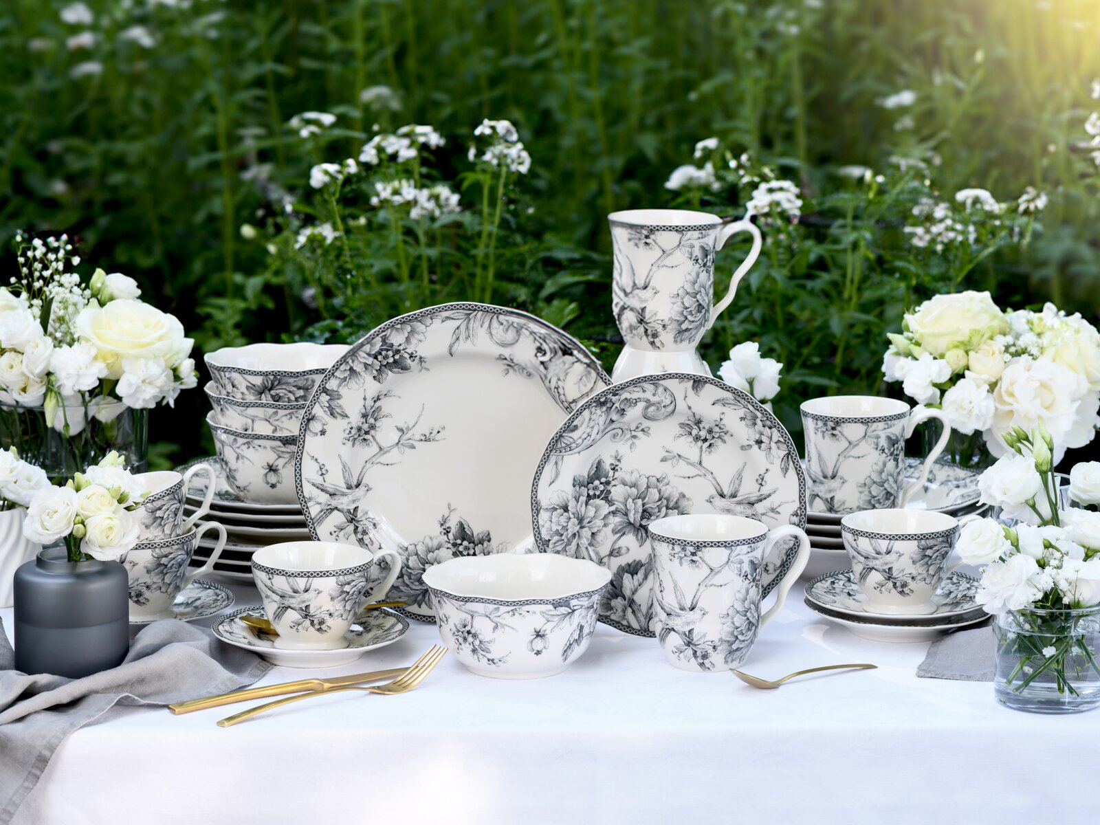 Schwarz-weißes Porzellan-Geschirrset mit floralen und Vogel-Motiven (Teller, Tassen, Schalen) auf gedecktem Tisch im Garten, Frontansicht mit leichter Aufsicht.