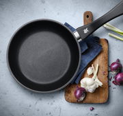 Pfanne aus einem 2-teiligen Set, Draufsicht, mit schwarzem Griff, auf einem Schneidebrett mit Zwiebeln und Knoblauch.