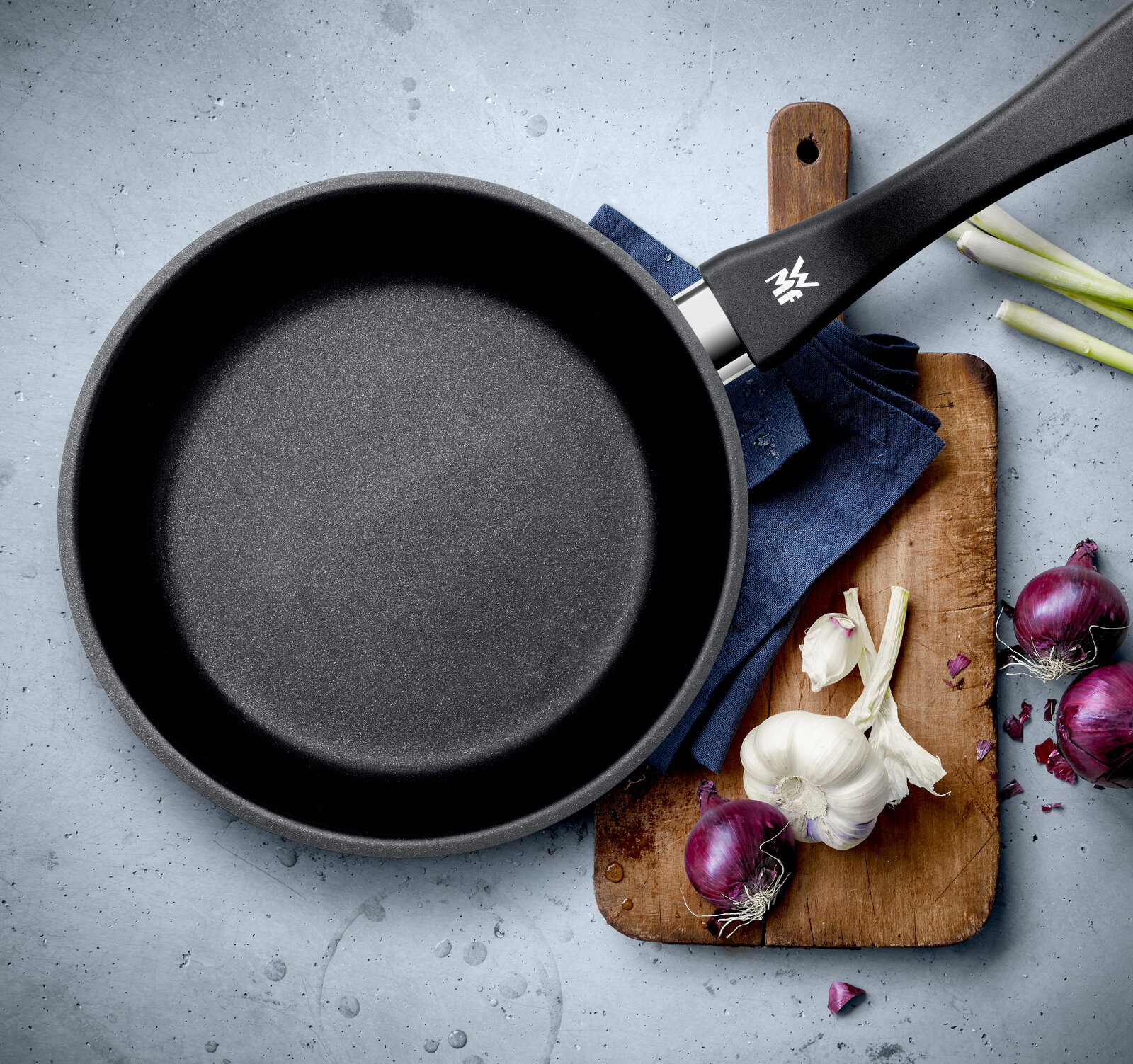 Pfanne aus einem 2-teiligen Set, Draufsicht, mit schwarzem Griff, auf einem Schneidebrett mit Zwiebeln und Knoblauch.