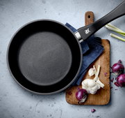 Pfanne aus einem 2-teiligen Set, Draufsicht, mit schwarzem Griff, auf einem Schneidebrett mit Zwiebeln und Knoblauch.