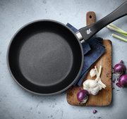 Pfanne aus einem 2-teiligen Set, Draufsicht, mit schwarzem Griff, auf einem Schneidebrett mit Zwiebeln und Knoblauch.