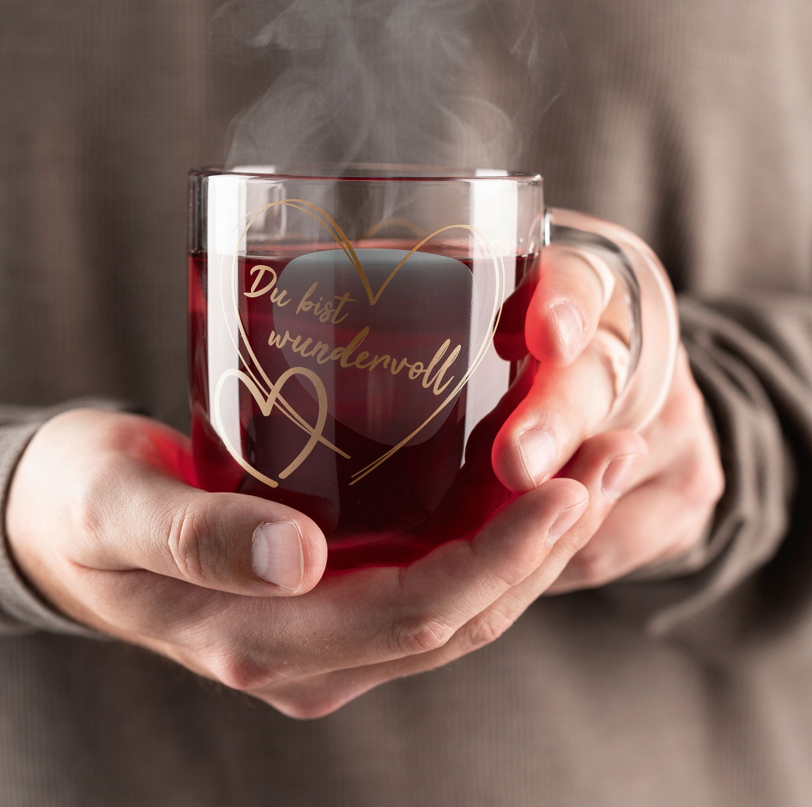 Leonardo Geschenktasse Du bist wundervoll EMOZIONE Glas-Tasse mit dampfendem Getränk, in Händen gehalten, mit der Aufschrift 'Du bist wundervoll' in goldener Schrift und Herzmotiv, aus der Frontalperspektive.