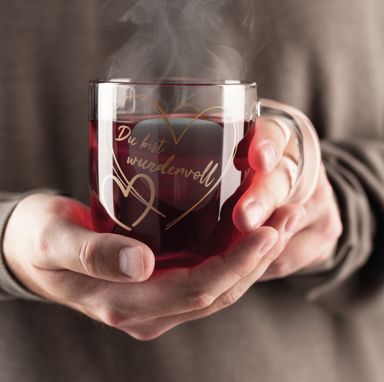 Glas-Tasse mit dampfendem Getränk, in Händen gehalten, mit der Aufschrift 'Du bist wundervoll' in goldener Schrift und Herzmotiv, aus der Frontalperspektive.