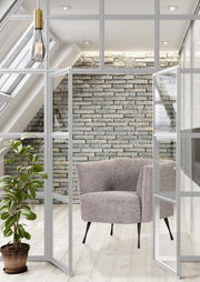 Moderner Sessel mit grauem Stoffbezug und schwarzen Beinen, in einem hellen Raum mit Glaswänden und Pflanzen, aus der Vorderansicht.