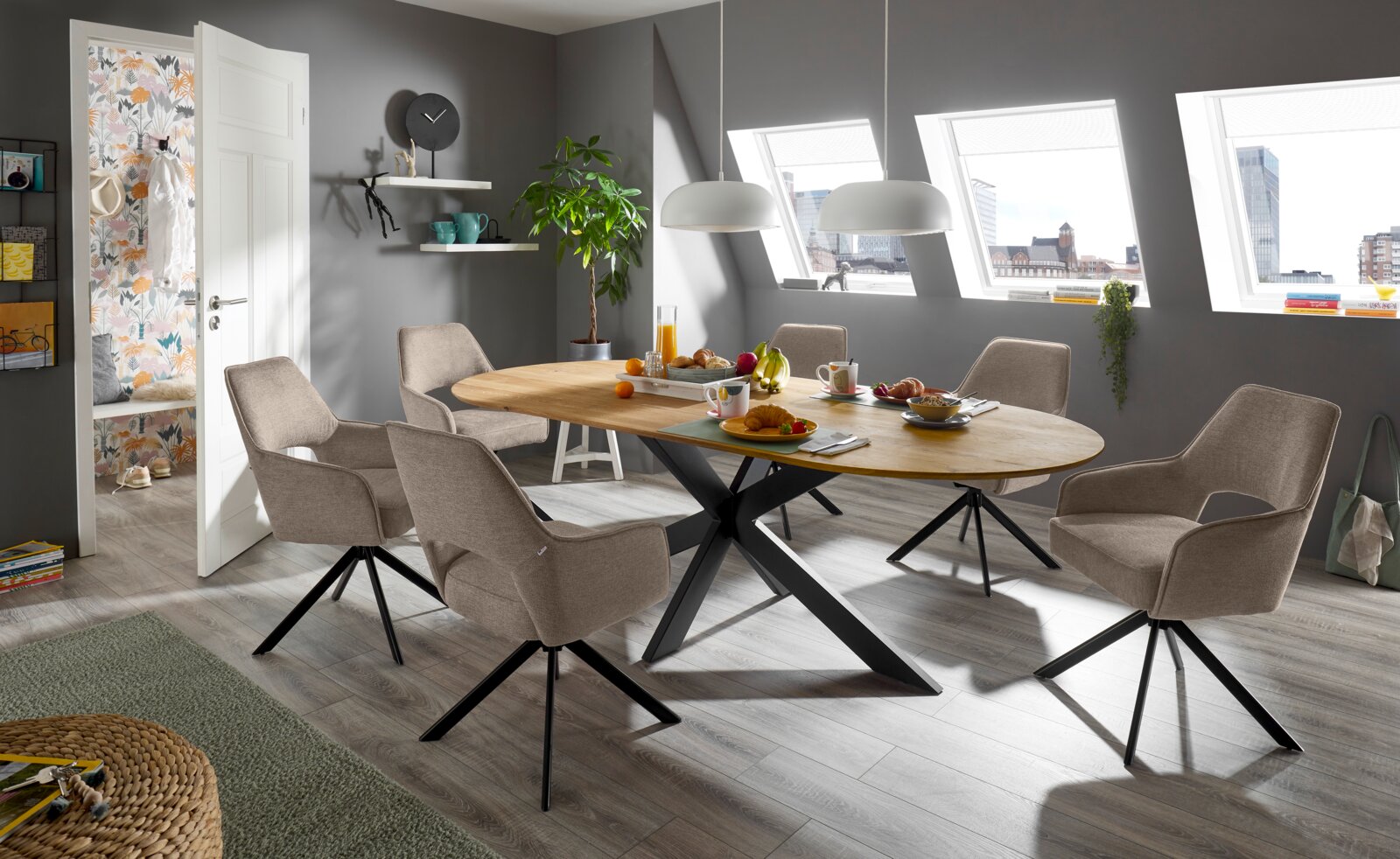 Moderner Esstisch mit ovaler Holzplatte und schwarzen Metallbeinen, umgeben von sechs gepolsterten Stühlen in einem stilvollen Esszimmer mit schrägen Dachfenstern, aus der Perspektive des Raumes betrachtet.