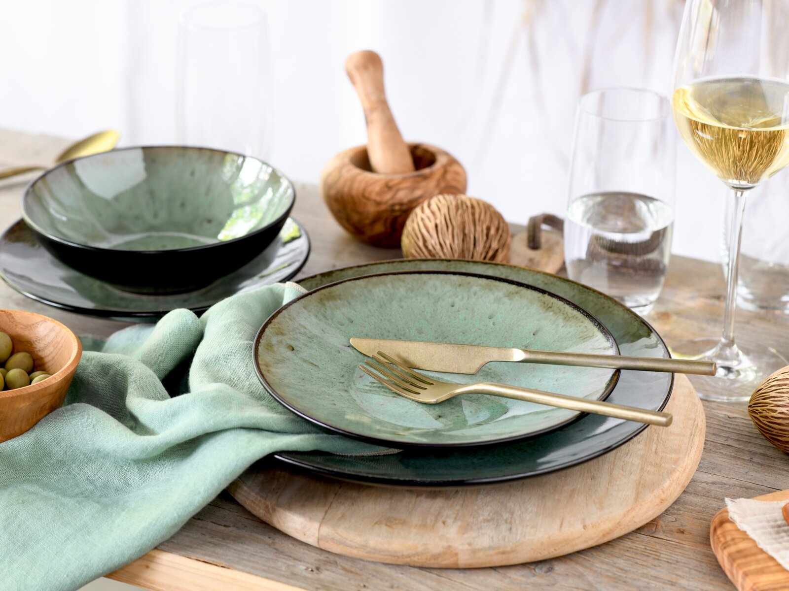 Rustikales Tisch-Arrangement mit grün glasiertem Keramikgeschirr (Teller und Schale), goldenem Besteck und Gläsern auf Holzbrett, fotografiert in leicht schräger Seitenansicht von vorne