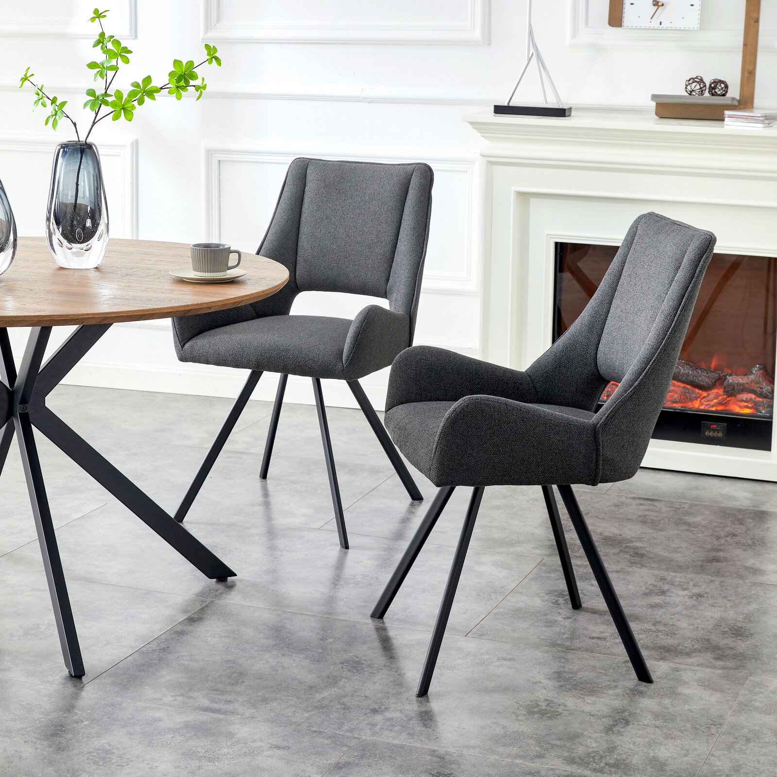 Zwei moderne Esszimmerstühle mit dunkelgrauem Stoffbezug, gepolsterter Sitzfläche und schwarzen Metallbeinen neben einem runden Holztisch, schräg von vorne in Innenraum-Szene fotografiert.