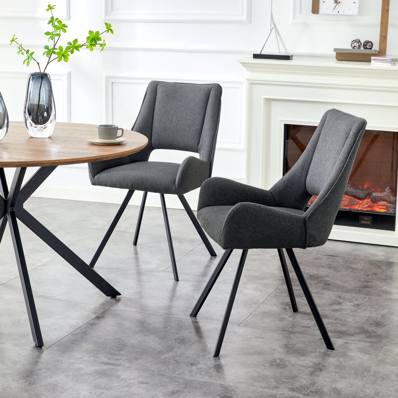 MID.YOU Stuhl 2er-Set HELEN Zwei moderne Esszimmerstühle mit dunkelgrauem Stoffbezug, gepolsterter Sitzfläche und schwarzen Metallbeinen neben einem runden Holztisch, schräg von vorne in Innenraum-Szene fotografiert.