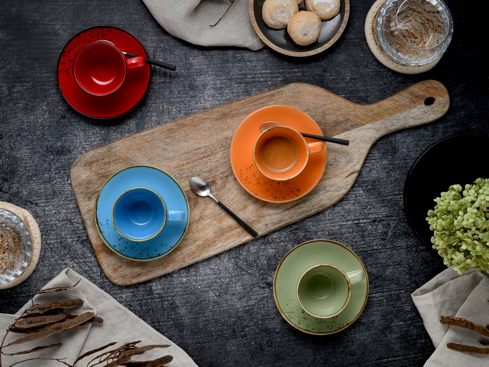 Draufsicht auf buntes Espressotassen- und Untertassen-Set (rot, orange, blau, grün) auf rustikalem Holzbrett mit Löffel, Gebäck und Deko auf dunklem Tisch