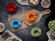 Draufsicht auf buntes Espressotassen- und Untertassen-Set (rot, orange, blau, grün) auf rustikalem Holzbrett mit Löffel, Gebäck und Deko auf dunklem Tisch