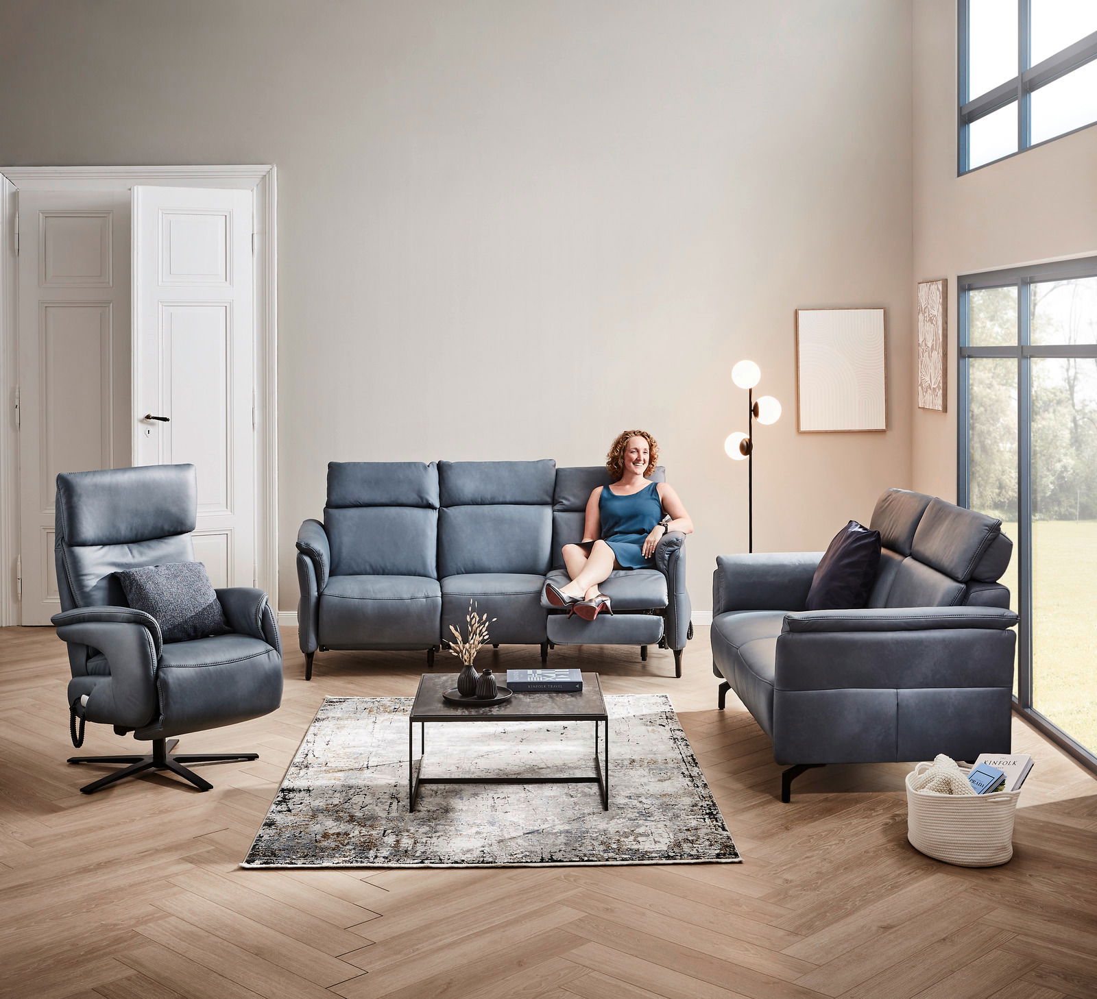 Wohnzimmeransicht mit einem blauen Rundsofa von Dieter Knoll, Modell VARO, in einer modernen Einrichtung mit einem Sessel, einem Couchtisch und einer Stehlampe. Eine Person sitzt entspannt auf dem Sofa. Perspektive: Frontalansicht.