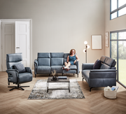 Wohnzimmeransicht mit einem blauen Rundsofa von Dieter Knoll, Modell VARO, in einer modernen Einrichtung mit einem Sessel, einem Couchtisch und einer Stehlampe. Eine Person sitzt entspannt auf dem Sofa. Perspektive: Frontalansicht.