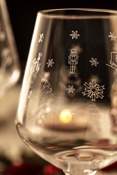Nahaufnahme eines Weißweinglases mit weihnachtlichen Gravuren wie Schneeflocken, Nussknacker und Tannenbaum, im Hintergrund unscharf ein weiteres Glas und Kerzenlicht.