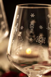 Nahaufnahme eines Weißweinglases mit weihnachtlichen Gravuren wie Schneeflocken, Nussknacker und Tannenbaum, im Hintergrund unscharf ein weiteres Glas und Kerzenlicht.