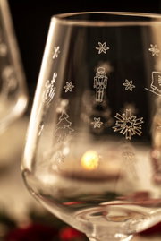 Close-up van een witwijnglas met kerstmotieven zoals sneeuwvlokken, notenkrakers en een kerstboom; op de achtergrond is vaag nog een glas en kaarslicht te zien.