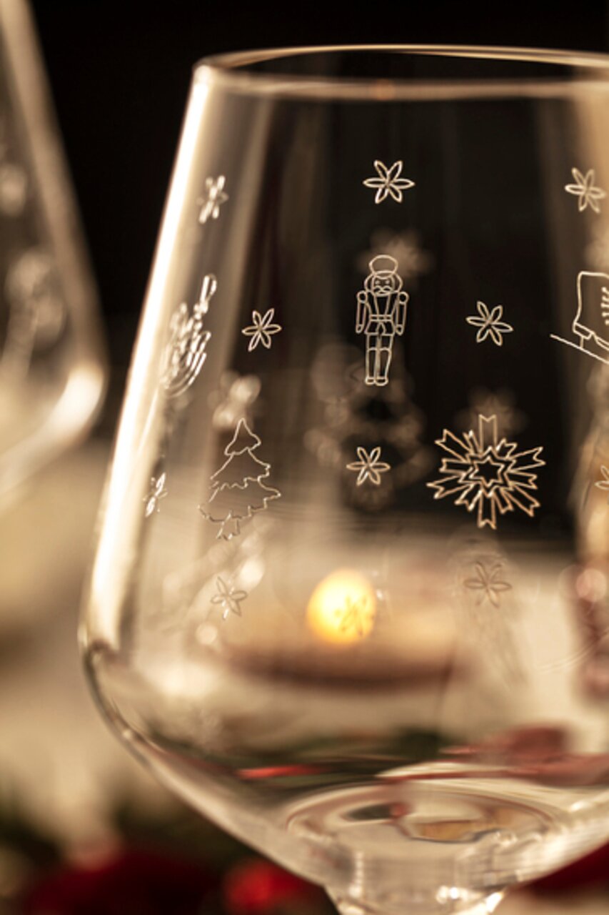 Close-up van een witwijnglas met kerstmotieven zoals sneeuwvlokken, notenkrakers en een kerstboom; op de achtergrond is vaag nog een glas en kaarslicht te zien.