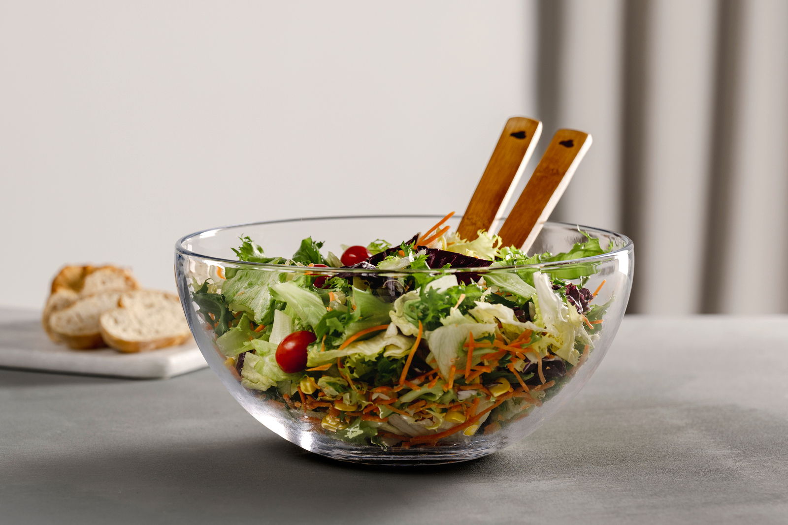 Glasklare Schale mit frischem Salat und Salatbesteck aus Holz, seitliche Perspektive