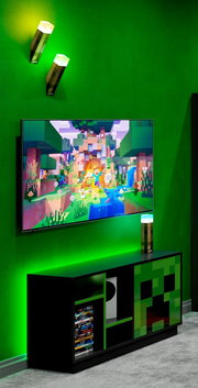 Schwarzes Gaming-TV-Lowboard mit grünem Pixel-Design (Creeper-Motiv) unter einem an der Wand montierten Fernseher, grüne LED-Ambientebeleuchtung und Minecraft-Fackel-Lampen; Perspektive: leicht schräg von vorne.