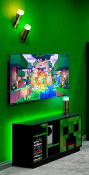 Schwarzes Gaming-TV-Lowboard mit grünem Pixel-Design (Creeper-Motiv) unter einem an der Wand montierten Fernseher, grüne LED-Ambientebeleuchtung und Minecraft-Fackel-Lampen; Perspektive: leicht schräg von vorne.