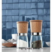 Twee kruidenmolens van hout en glas, gevuld met zout en peper, op een keukenblad voor een blauwe tegelwand, zijaanzicht.