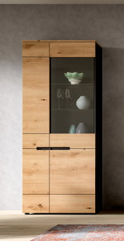 Hoher Vitrinenschrank aus hellem Holz mit Glastür und schwarzen Griffen, frontale Ansicht; im Inneren stehen Gläser und dekorative Vasen.