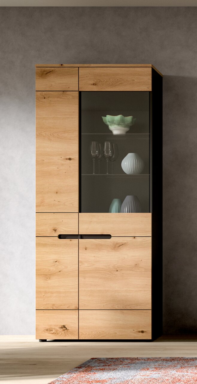 Hoher Vitrinenschrank aus hellem Holz mit Glastür und schwarzen Griffen, frontale Ansicht; im Inneren stehen Gläser und dekorative Vasen.